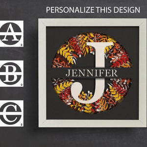 May include: A framed paper art piece with a white frame. The design features a large white letter "J" and the name "Jennifer", set against a backdrop of autumn leaves in red, orange, and yellow. The text "PERSONALIZE THIS DESIGN" is at the top.