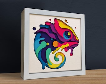 Peut inclure: Une œuvre d'art en papier découpé colorée représentant un caméléon aux couleurs vives violettes, bleues, vertes, jaunes et rouges. Le caméléon est encadré dans un cadre blanc.