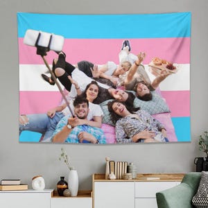 May include: A wall tapestry featuring the transgender pride flag design, with a group of people lying on a bed, taking a selfie. The flag has horizontal stripes of light blue, pink, and white. The people are smiling and waving.
