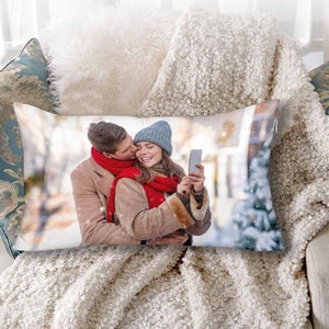 K&ouml;nnte beinhalten: Ein wei&szlig;es Kissen mit einem gedruckten Bild eines Paares in einer verschneiten Umgebung. Das Paar tr&auml;gt Winterkleidung und h&auml;lt ein Telefon. Das Bild ist auf der Vorderseite des Kissens gedruckt.