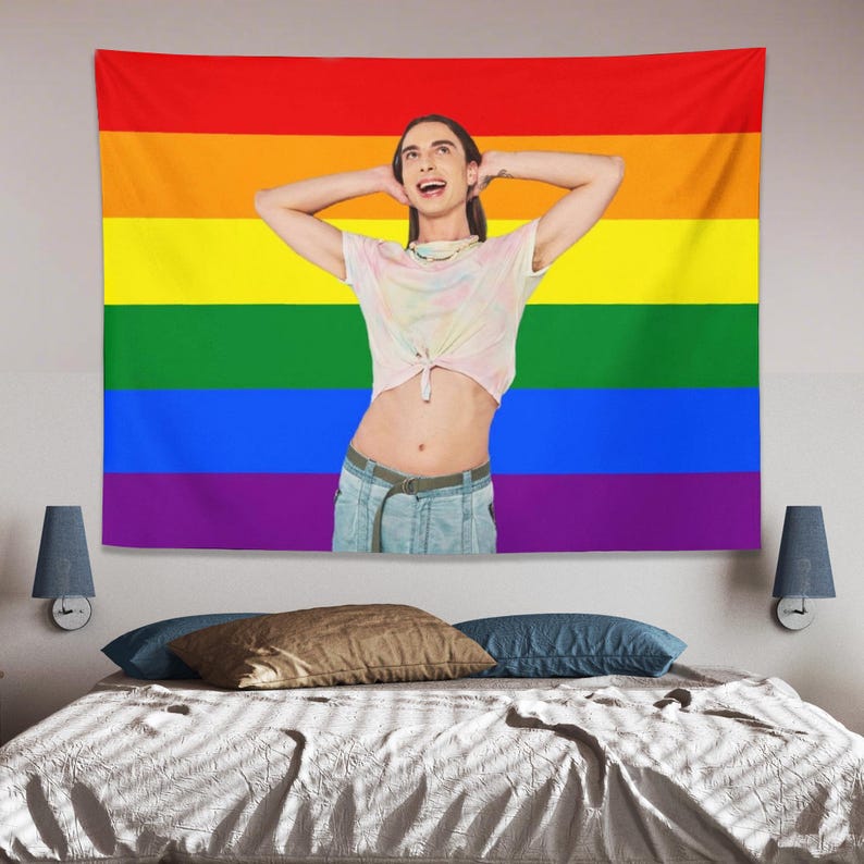 May include: A large rainbow flag tapestry with horizontal stripes of red, orange, yellow, green, blue, and purple. A person is in front of the flag, wearing a tie-dye shirt and denim shorts. The person is smiling with their arms behind their head.