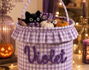 May include: A cylindrical fabric basket featuring a purple and white gingham pattern with a ruffled trim and a matching handle. The name Violet is embroidered in purple yarn on the front. The basket is filled with small pumpkins and a black cat toy.
