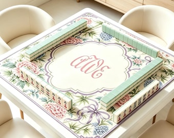 May include: A square mahjong mat featuring a floral pattern with pink peonies, blue hydrangeas, and green bamboo leaves. The center displays the monogram "aWe" in a decorative frame with a purple border. Four sets of light green mahjong tiles are arranged.