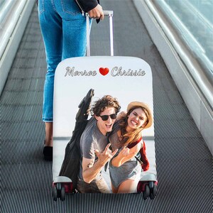 以下が含まれることがあります： 赤いハートと「Monroe &hearts; Chrissie」の文字が書かれた白いスーツケースカバー。カバーには、笑顔でロックンロールのハンドジェスチャーをしているカップルの写真が写っています。