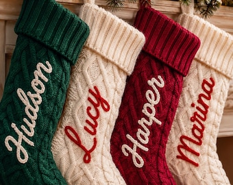 May include: Four cable-knit Christmas stockings hanging in a row. The first is dark green with the white embroidered name Hudson. The second is cream with the red embroidered name Lucy. The third is dark red with the white name Harper. The fourth is cream with the red name Marina.