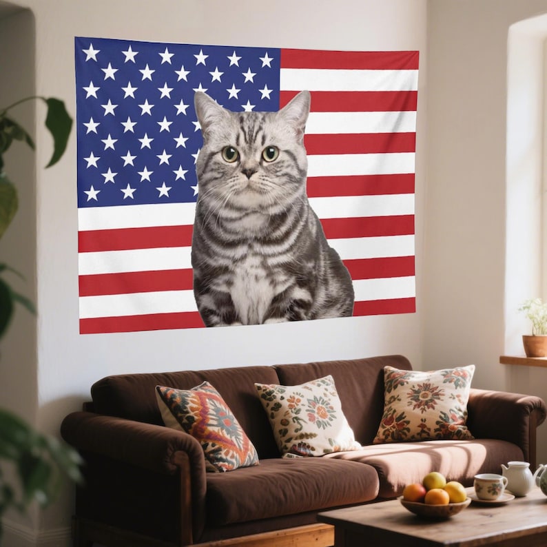 May include: A gray tabby cat sits in front of an American flag tapestry. The cat has green eyes and a striped coat. The flag is red, white, and blue, with stars and stripes. A brown sofa with patterned pillows is in the foreground.