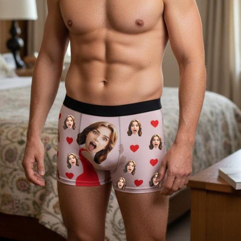 May include: Pink boxer briefs with a black waistband, featuring a repeating pattern of red hearts and faces. The faces are of a woman with an open mouth. The woman is wearing a red dress.