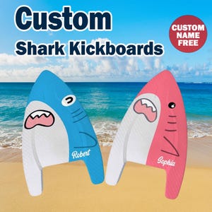 Puede incluir: Dos tablas de natación con forma de tiburón, una azul y otra rosa, con vientre blanco, cara de tiburón de dibujos animados y un nombre personalizado impreso en blanco. El texto Custom Shark Kickboards y un círculo rojo CUSTOM NAME FREE.