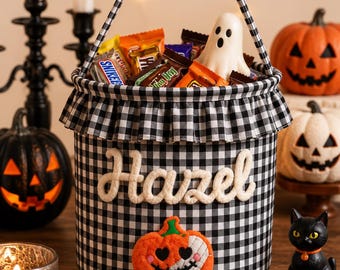 May include: A black and white gingham fabric bucket with a ruffled top edge and a matching handle. The front features the name Hazel in white script lettering above a plush orange pumpkin patch with a stitched face and heart-shaped eyes.