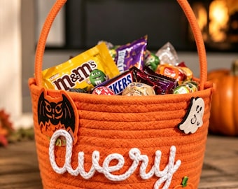 May include: An orange rope basket with a handle, featuring the name Avery in white rope script. The basket is decorated with felt appliques of a black bat, a white ghost, and an orange jack-o'-lantern, and is filled with various candy wrappers.