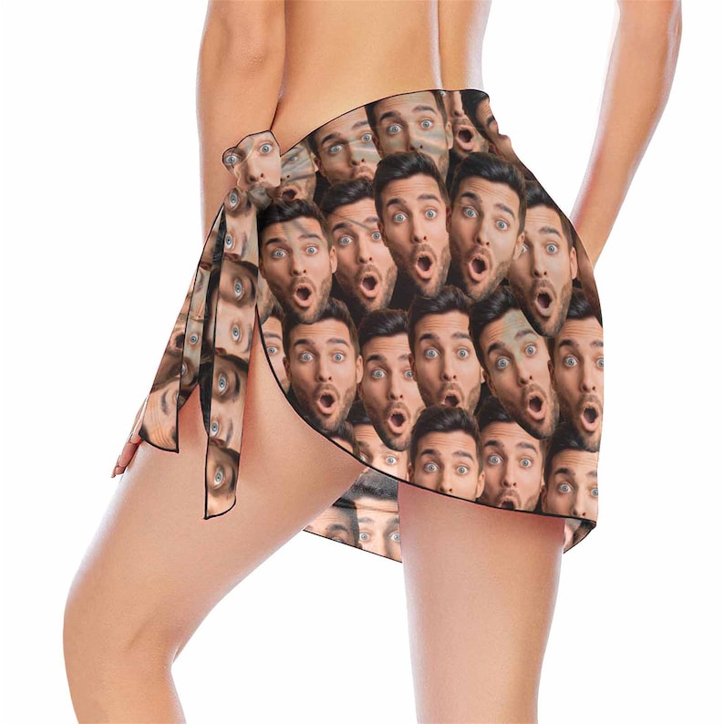 May include: A black sarong with a repeating pattern of a man's face with a surprised expression. The sarong is tied around the waist and has a long tie that hangs down the side.