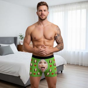 May include: Green boxer briefs with a black waistband, featuring a repeating pattern of red hearts and a woman's face with a zipper design. The briefs are worn by a person.