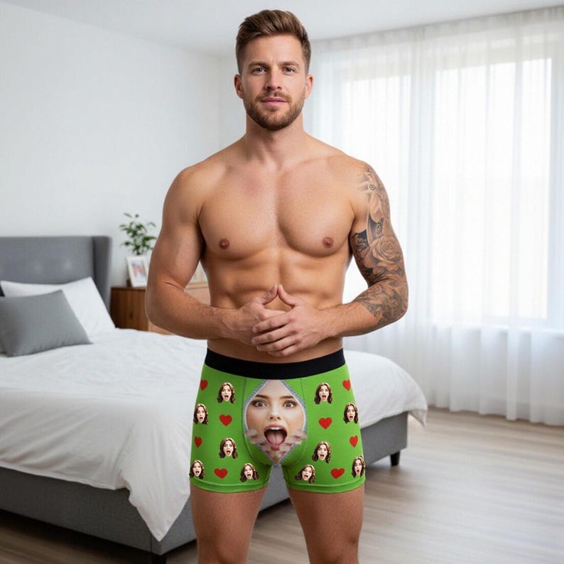 May include: Green boxer briefs with a black waistband, featuring a repeating pattern of red hearts and a woman's face with a zipper design. The briefs are worn by a person.