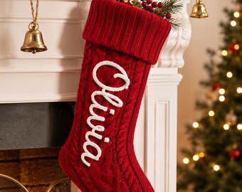 May include: A deep red cable-knit Christmas stocking featuring a folded cuff at the top and the name Olivia embroidered in white script lettering on the front. The stocking includes a matching red fabric loop for hanging and a textured pattern.