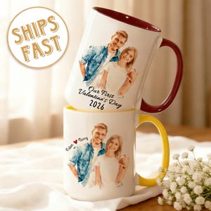 May include: Two white ceramic mugs stacked, each featuring a couple's portrait. One mug has a burgundy handle and rim, the other a yellow handle and rim. Text on the mugs reads "Our First Valentine's Day 2026" and names with a heart symbol.