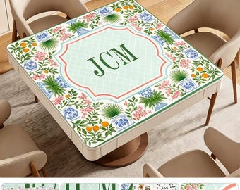 May include: A square mahjong mat featuring a light green grid pattern with a central monogram JCM in dark green. The border displays a botanical print of orange trees, pink flowers, and blue and white patterned vases on a white background.