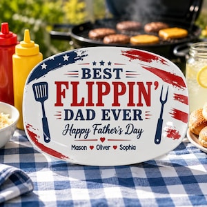 Puede incluir: Plato ovalado blanco con diseño rojo, blanco y azul. El texto dice "BEST FLIPPIN' DAD EVER Happy Father's Day" con nombres debajo. Una espátula y un tenedor están a cada lado del texto. El plato está sobre un mantel a cuadros azul y blanco.