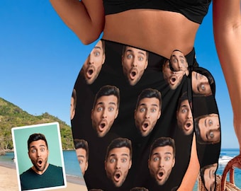 May include: A black beach wrap featuring a repeating pattern of a man's face with a surprised expression, with an open mouth and wide eyes. The wrap is tied at the side, and the fabric displays the same facial print throughout the entire garment.