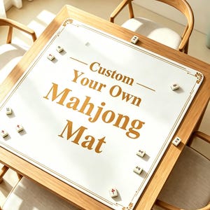 May include: A square white mahjong mat with gold decorative corner accents and gold text that reads Custom Your Own Mahjong Mat. The mat is placed on a wooden table and is scattered with several small white mahjong tiles featuring red and black symbols.