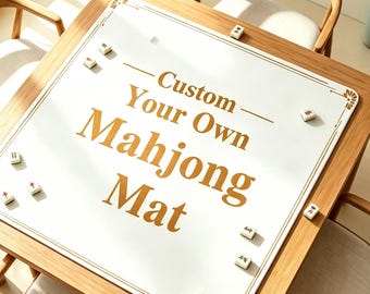 May include: A square white mahjong mat with gold decorative corner accents and gold text that reads Custom Your Own Mahjong Mat. The mat is placed on a wooden table and is scattered with several small white mahjong tiles featuring red and black symbols.