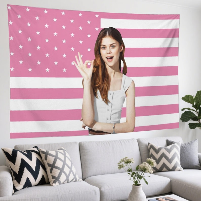 May include: Pink and white American flag tapestry hanging on a wall. A woman is standing in front of the tapestry, making an 'okay' hand gesture. A white couch with decorative pillows and a vase of flowers are in front of the wall.