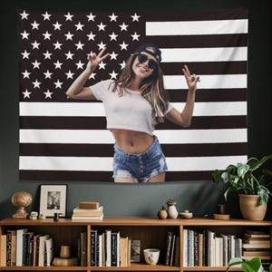 May include: A black and white American flag tapestry with a person in the center. The person is wearing a white crop top, denim shorts, sunglasses, and a baseball cap. The person is making a peace sign with both hands.