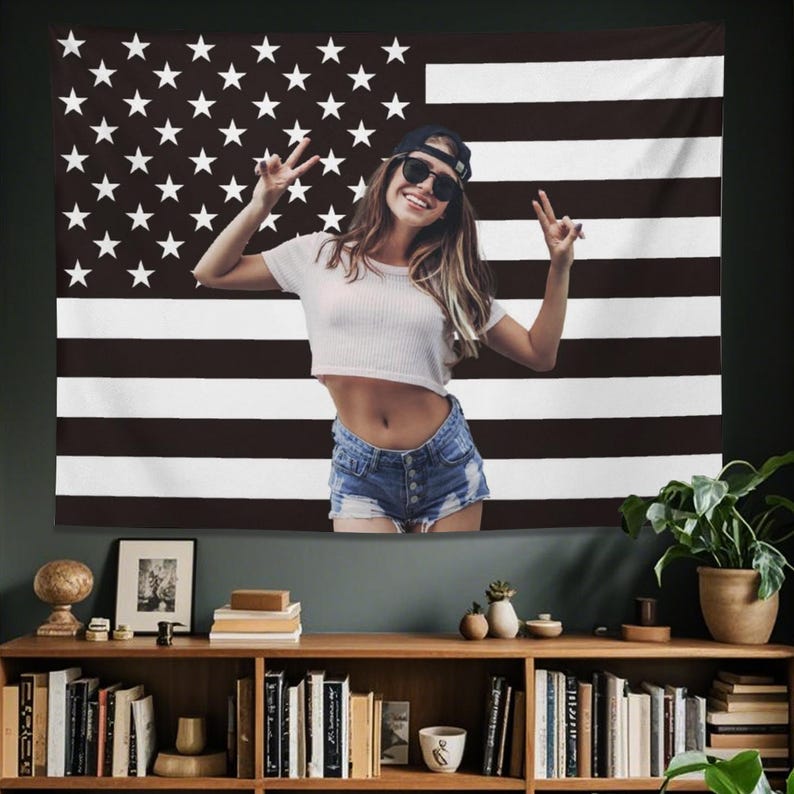 May include: A black and white American flag tapestry with a person in the center. The person is wearing a white crop top, denim shorts, sunglasses, and a baseball cap. The person is making a peace sign with both hands.