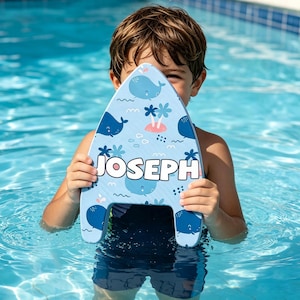 May include: A light blue foam kickboard featuring a repeating pattern of dark blue whales, small pink palm trees, and white bubbles. The name JOSEPH is printed in large, bold white capital letters across the center of the aquatic swimming board.