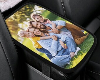 May include: A rectangular car center console armrest cover featuring a printed photograph of a family of four sitting together on grass. The cover has a black border and is secured to the black leather console with an elastic strap.
