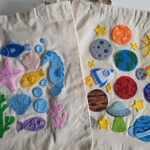 May include: Two beige canvas tote bags with colourful felt appliqués. One bag features sea creatures like a seahorse, fish, and starfish. The other bag has planets, stars, and a rocket ship. Both bags have cream-coloured handles.