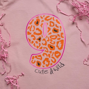 Könnte beinhalten: Hellrosa T-Shirt mit einer großen Zahl 9 mit einem orangefarbenen Leopardenmuster und einem leuchtend rosa Umriss. Die Worte "Cute & Wild" sind in schwarzer Schrift unter der Zahl geschrieben. Rosa Papierschnipsel sind um das Shirt verstreut.