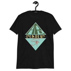 Könnte beinhalten: Schwarzes T-Shirt mit einem geometrischen Design in Grün und Türkis. Das Design zeigt die Buchstaben "ARB", den Schriftzug "EYE IN THE SKY" und ein Augensymbol. Das T-Shirt hängt an einem schwarzen Kleiderbügel.