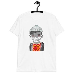 Puede incluir: Camiseta blanca con una ilustración de dibujos animados de una persona que lleva un sombrero y una mascarilla. La mascarilla dice "IT'S OK TO BE HUMAN". La camiseta de la persona tiene un diseño de corazón. La camiseta está colgada en una percha negra.