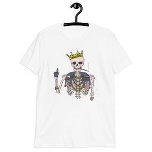 Puede incluir: Camiseta blanca con un gráfico de un esqueleto que lleva una corona dorada, cadenas doradas y un cigarrillo. El esqueleto está haciendo un signo de paz con una mano.