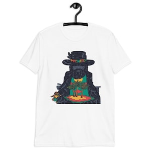 Puede incluir: Camiseta blanca con un gráfico de una figura barbuda que lleva un sombrero con la palabra "IMAGINATION". La figura tiene una escena de isla tropical en el pecho. La camiseta está colgada en una percha negra.