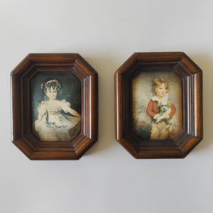 May include: Two octagonal wooden frames containing portrait prints. The left frame holds a print of a person in a white dress with a dark headpiece. The right frame holds a print of a person in a red jacket holding a small white dog.