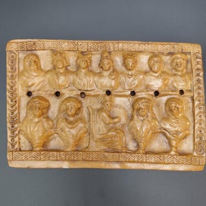 May include: A rectangular tan-colored plaque featuring a relief carving of twelve human figures arranged in two rows. The border is decorated with a repeating geometric pattern. A horizontal line of six small circular holes runs across the center.