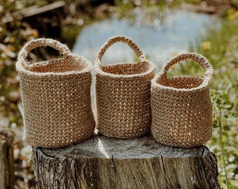 May include: Three cylindrical hanging baskets made of natural tan jute twine, each featuring a single arched handle at the top. The baskets are arranged in a row, showing a graduated size progression from the largest on the left to the smallest on the right.