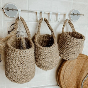May include: Three natural fibre hanging baskets with rope handles. The baskets are made of a woven material and are hanging from a metal rod with hooks. The baskets are against a tiled wall.