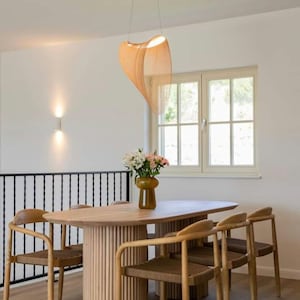 Op de afbeelding: Een houten eettafel met een lichte houtkleur en een unieke ovale vorm. De tafel heeft een geflute basis en wordt omringd door zes stoelen met gevlochten zittingen. Een boeket bloemen in een gele vaas staat op de tafel.