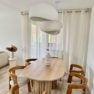 Könnte beinhalten: Ein ovaler Esstisch aus hellem Holz mit einem einzigartigen Lattengestell und sechs Stühlen. Der Tisch ist mit einer weißen Keramikvase mit drei übereinander gestapelten weißen Keramikschalen gedeckt. Der Raum hat weiße Wände und weiße, durchsichtige Vorhänge.