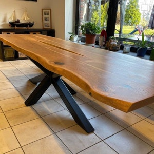 Op de afbeelding: Een grote, rustieke houten eettafel met een live edge blad en een zwarte metalen X-vormige basis. De tafel is gemaakt van één stuk hout en heeft een natuurlijke, organische vorm.