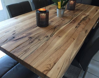 Op de afbeelding: Een grote, rechthoekige houten eettafel met een natuurlijke houtnerf afwerking. De tafel is omringd door donkergrijze stoelen. Decoratieve items, waaronder een kaars in een zwarte metalen lantaarn en een kleine potplant, staan op de tafel.