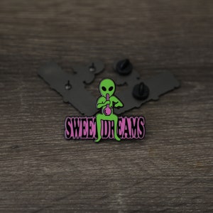 Könnte beinhalten: Schwarzer Emaille-Pin mit einer grünen Alien-Karikatur, die eine Bong raucht, und dem Text "SWEET DREAMS" in rosa Buchstaben.