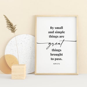 May include: A framed print with a white background and black text that reads "By small and simple things are great things brought to pass. ALMA 37:6". The print is sitting on a white surface with a wooden block and two round objects.