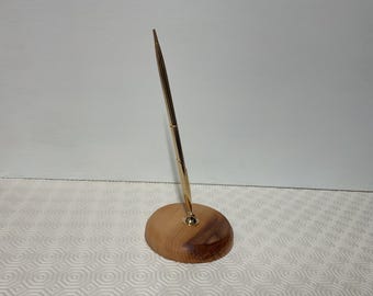 May include: A gold-colored pen stands upright in a wooden pen holder. The pen has a sleek, modern design, and the holder is a natural wood color. The pen and holder are set against a white background.