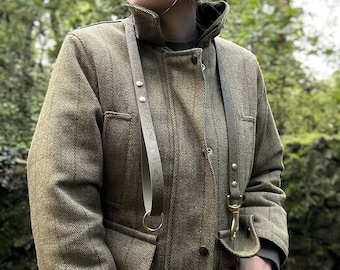 May include: A person wearing a green tweed jacket with two leather straps and brass hardware. The straps are attached to a ring and a hook.