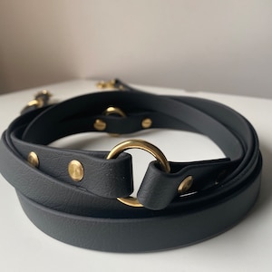 May include: A black leather belt with a gold-coloured metal ring and rivets. The belt is coiled, showcasing its design and hardware. The hardware includes a gold-coloured metal ring and rivets.