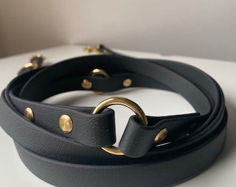 May include: A black leather belt with a gold-colored metal ring and rivets. The belt is coiled, showcasing its design and hardware. The hardware includes a gold-colored metal ring and rivets.