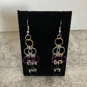 Könnte beinhalten: Ein Paar Kettenhemd-Ohrhänger mit einer vertikalen Anordnung ineinandergreifender Metallringe. Das Design besteht aus silbernen, goldenen, violetten und schwarzen Ringen, die an einem silberfarbenen Ohrhaken befestigt sind.